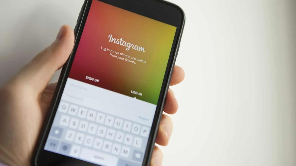 Hand holding a black smartphone displaying the Instagram login screen with a colorful gradient background. White keyboard visible.