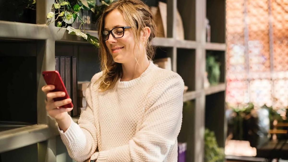 Mujer con gafas sonriendo mientras mira su teléfono en una tienda con estanterías de madera