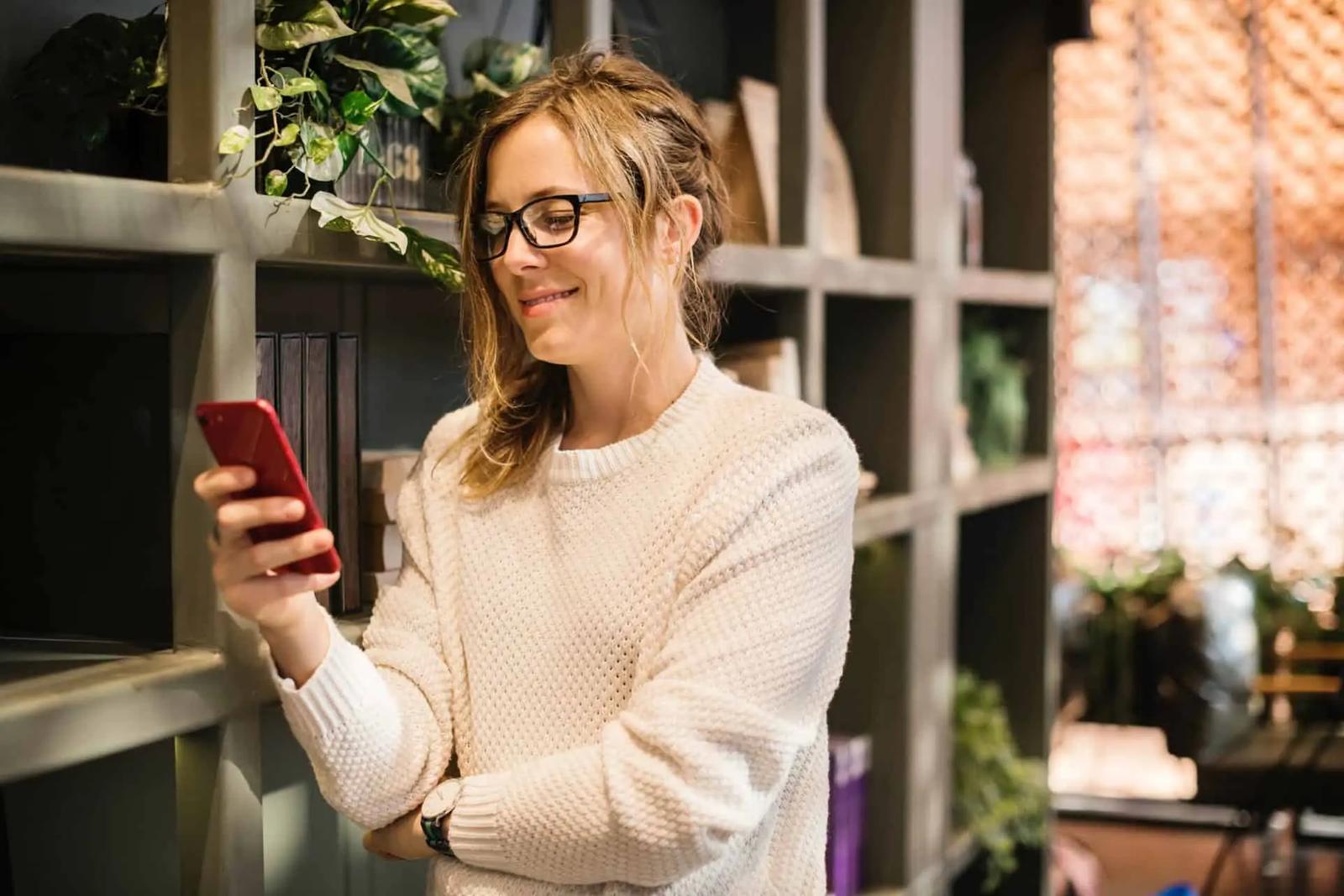 Mujer con gafas sonriendo mientras mira su teléfono en una tienda con estanterías de madera