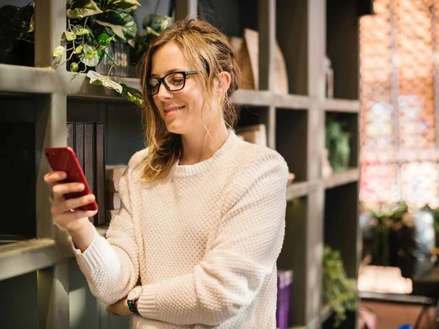Mujer con gafas sonriendo mientras mira su teléfono en una tienda con estanterías de madera