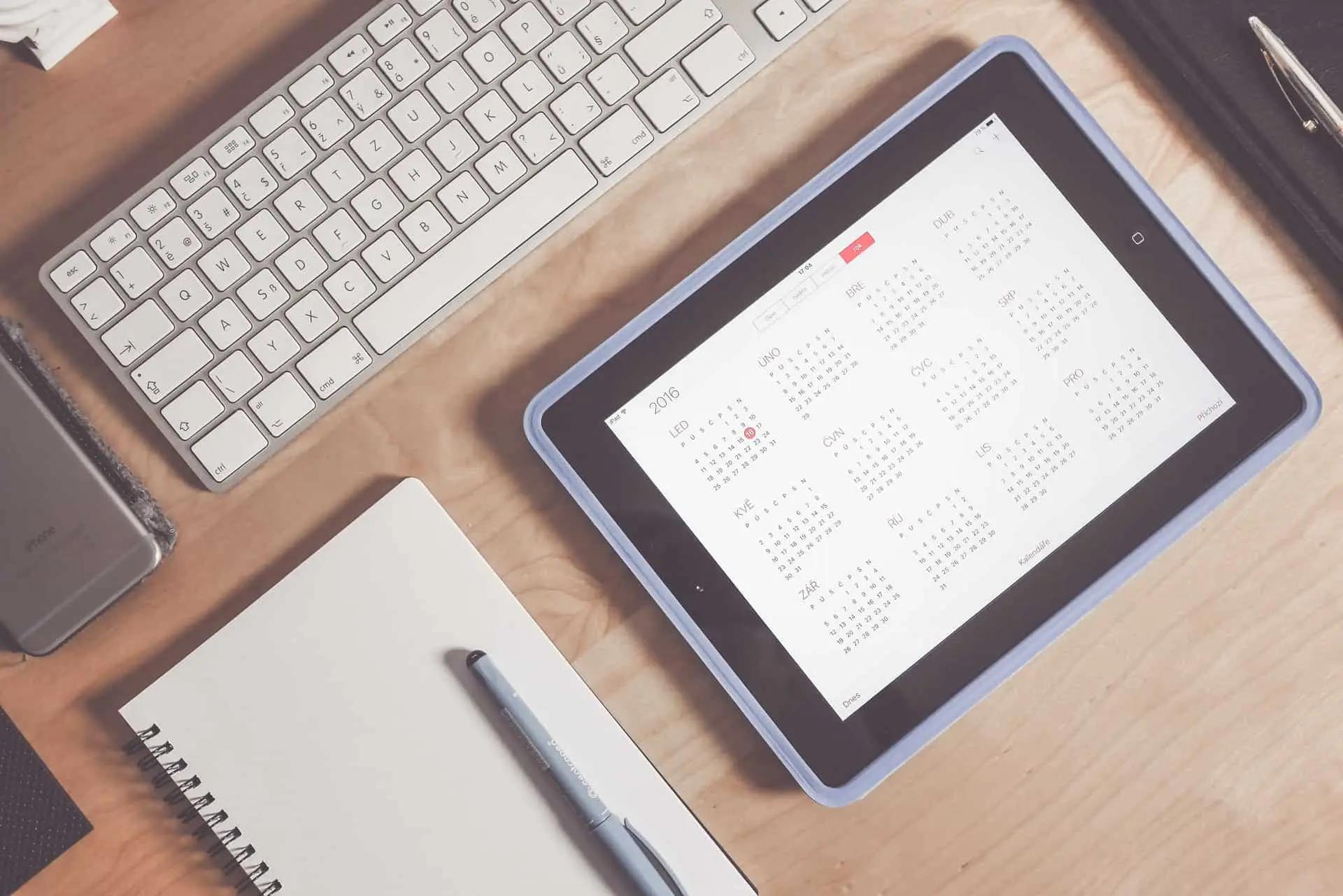 Overhead view of a desk with a keyboard, notebook, phone and a tablet showing a yearly calendar app
