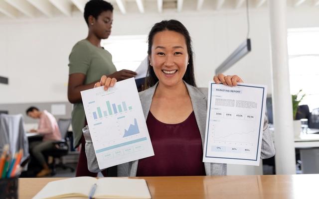 Smiling woman holding two marketing reports with charts. Two other people are in the background of a bright office.