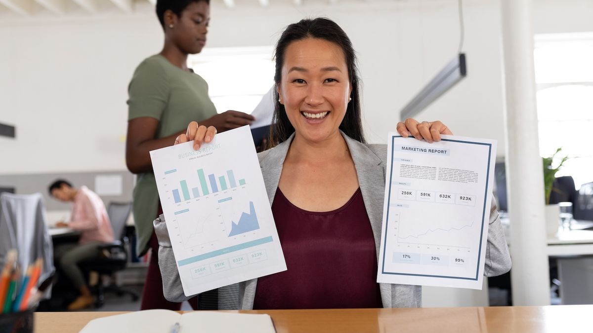 Smiling woman holding two marketing reports with charts. Two other people are in the background of a bright office.