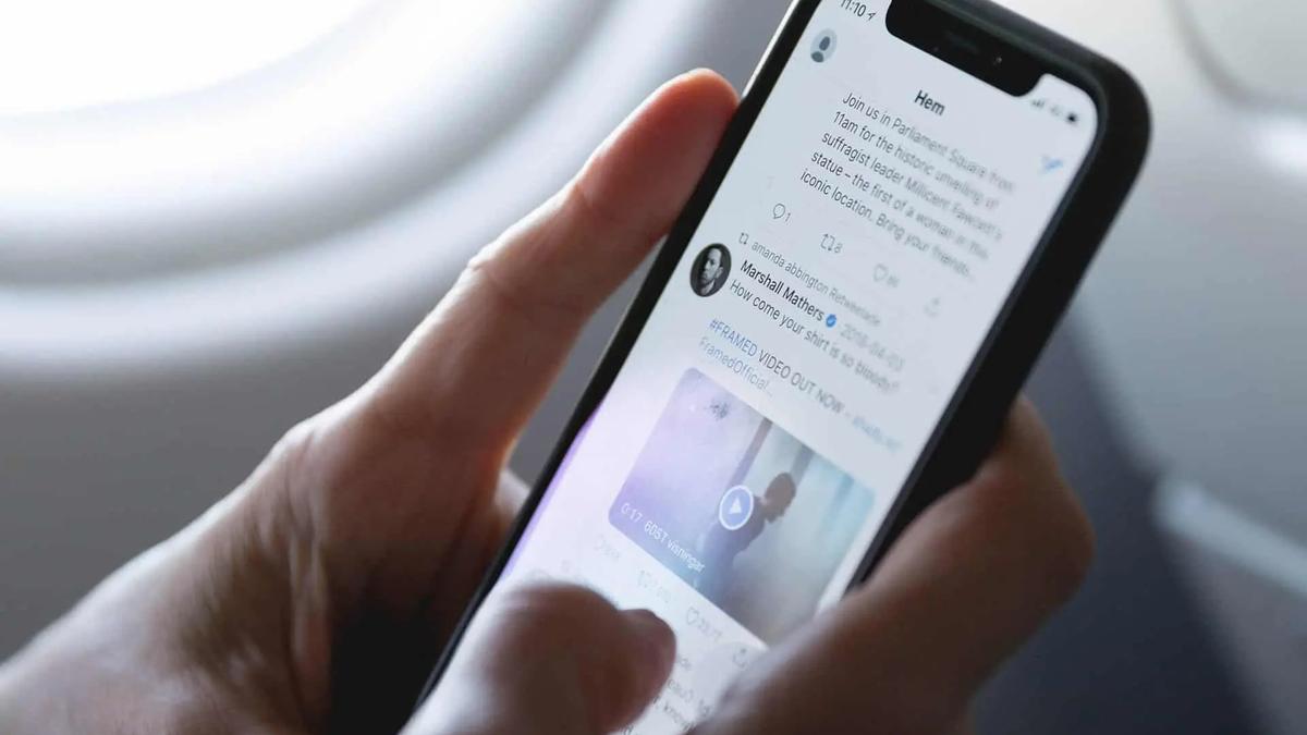 Una mano desplaza el feed de una red social en un smartphone junto a la ventanilla de un avion