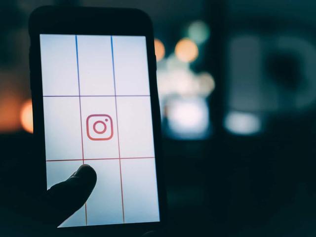 Una mano sostiene un teléfono con el logo de Instagram sobre una pantalla en cuadrícula, con luces difuminadas de fondo