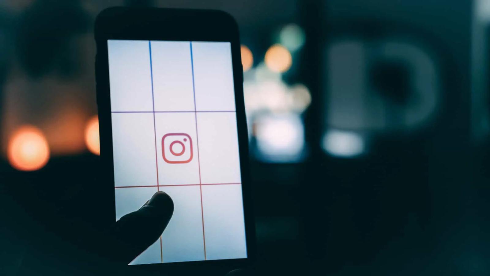 A hand holds a phone showing the Instagram logo on a grid screen, with blurred lights behind