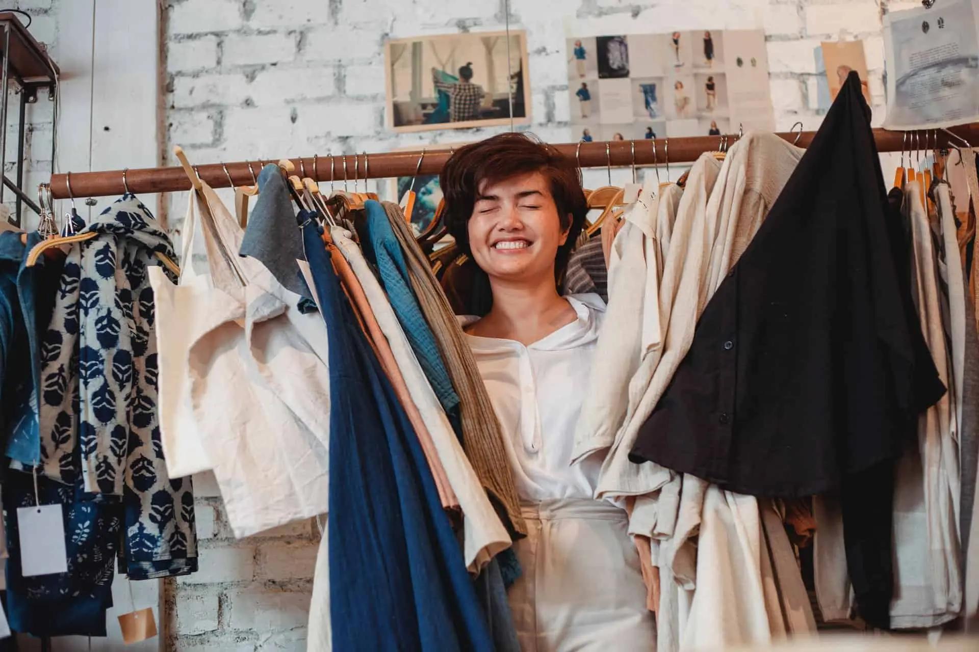 A smiling woman stands among clothing racks in a shop, surrounded by hanging garments and design photos.