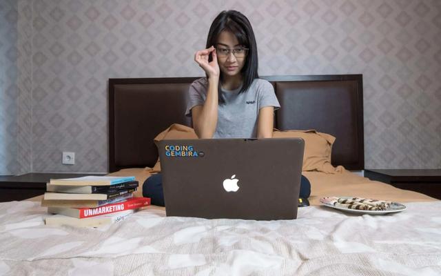 Woman in glasses and gray shirt sitting on a bed with a laptop, books, and a plate of pastries. Calm mood.