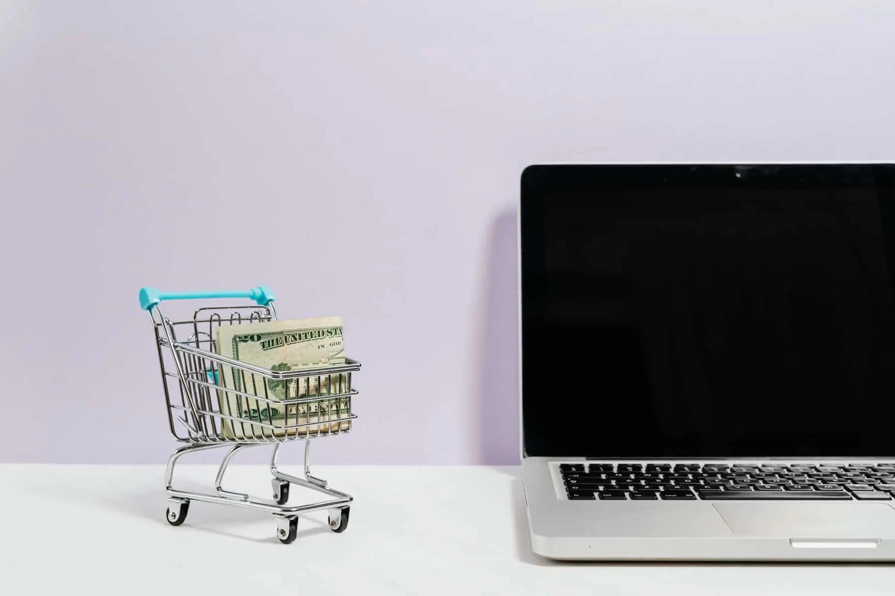 Un carrito de compras en miniatura con billetes doblados junto a una laptop abierta sobre un escritorio