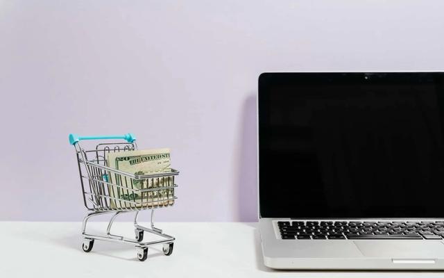 A miniature shopping cart holding folded banknotes next to an open laptop on a desk