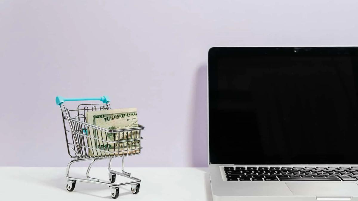 Un carrito de compras en miniatura con billetes doblados junto a una laptop abierta sobre un escritorio