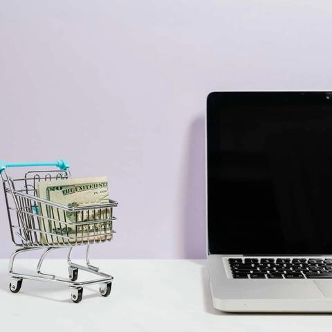 Un carrito de compras en miniatura con billetes doblados junto a una laptop abierta sobre un escritorio