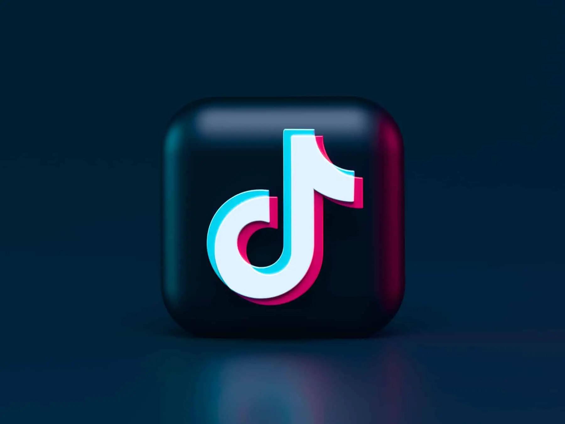 3D TikTok logo, white with pink and blue shadows, on a dark blue background with reflections. Modern, clean mood.