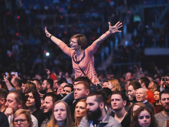Woman with short hair, glasses, and a striped shirt on someone's shoulders in a large, dark crowd, arms raised.