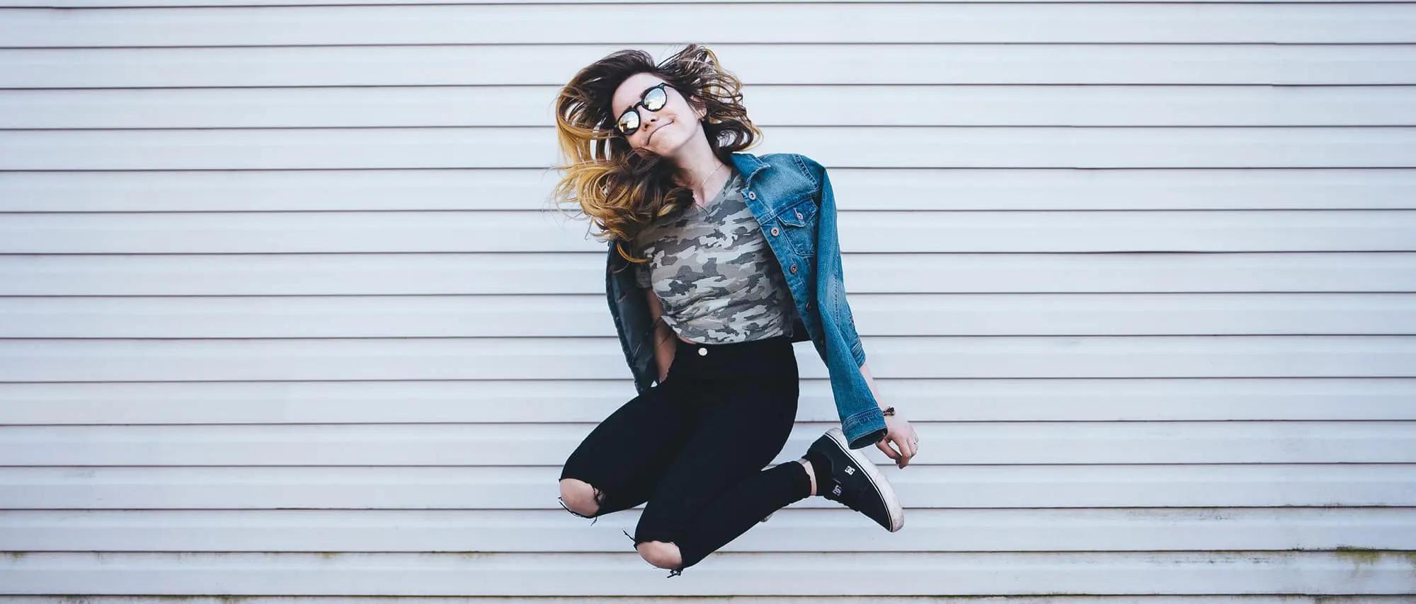 Una joven con gafas de sol y chaqueta vaquera saltando en el aire frente a una pared de metal corrugado