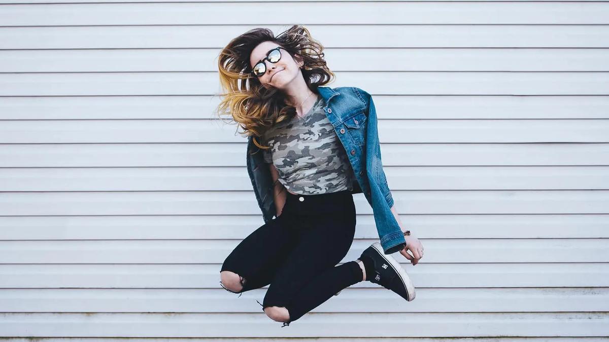 A young woman in sunglasses and a denim jacket jumping mid-air in front of a corrugated metal wall