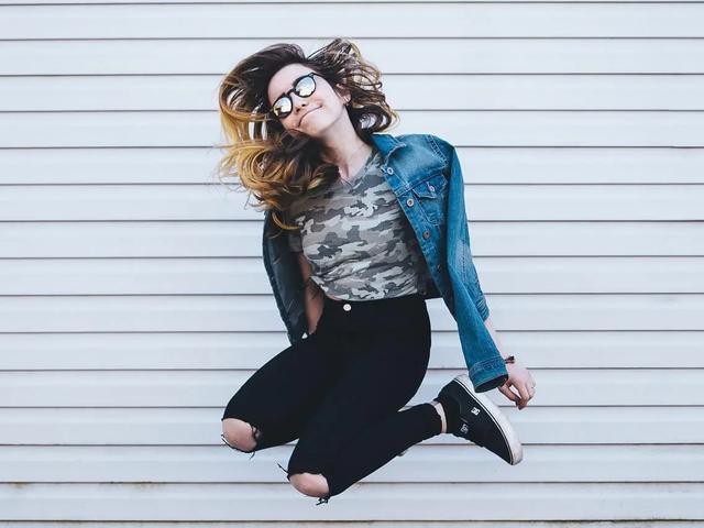A young woman in sunglasses and a denim jacket jumping mid-air in front of a corrugated metal wall
