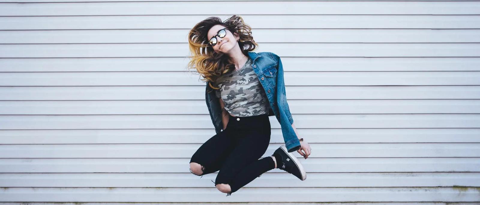 A young woman in sunglasses and a denim jacket jumping mid-air in front of a corrugated metal wall