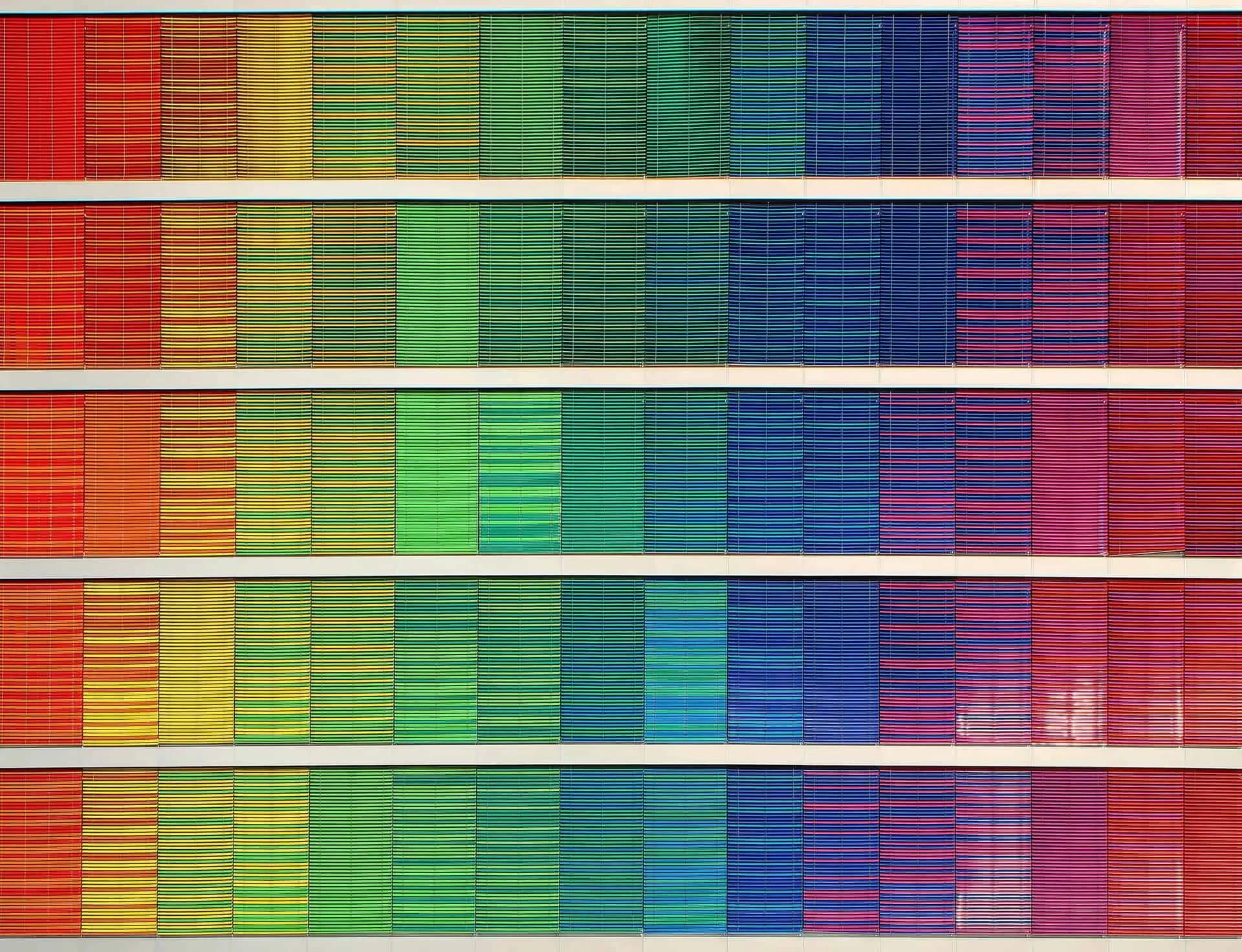 Rows of colourful window blinds forming a rainbow gradient pattern on a building facade