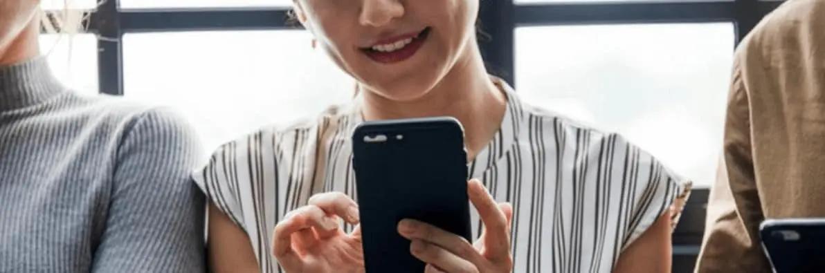 A smiling woman in a striped top holding and looking at a smartphone, seated between others
