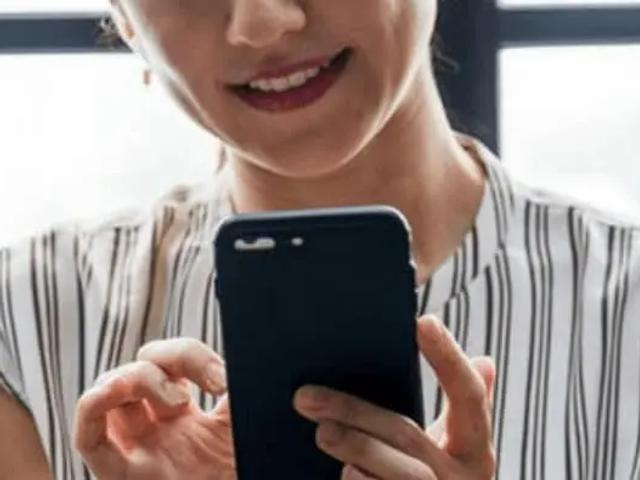 Una mujer sonriente con blusa de rayas sostiene y mira un smartphone, sentada entre otras personas