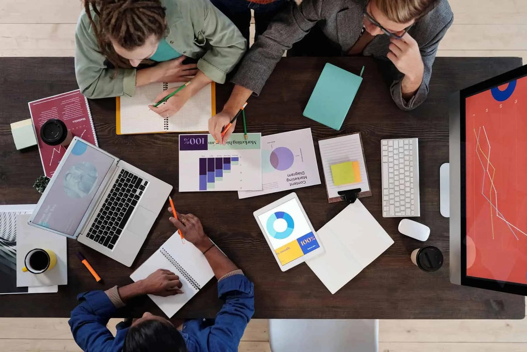 Overhead view of a team at a desk reviewing marketing charts, laptops and a tablet with graphs
