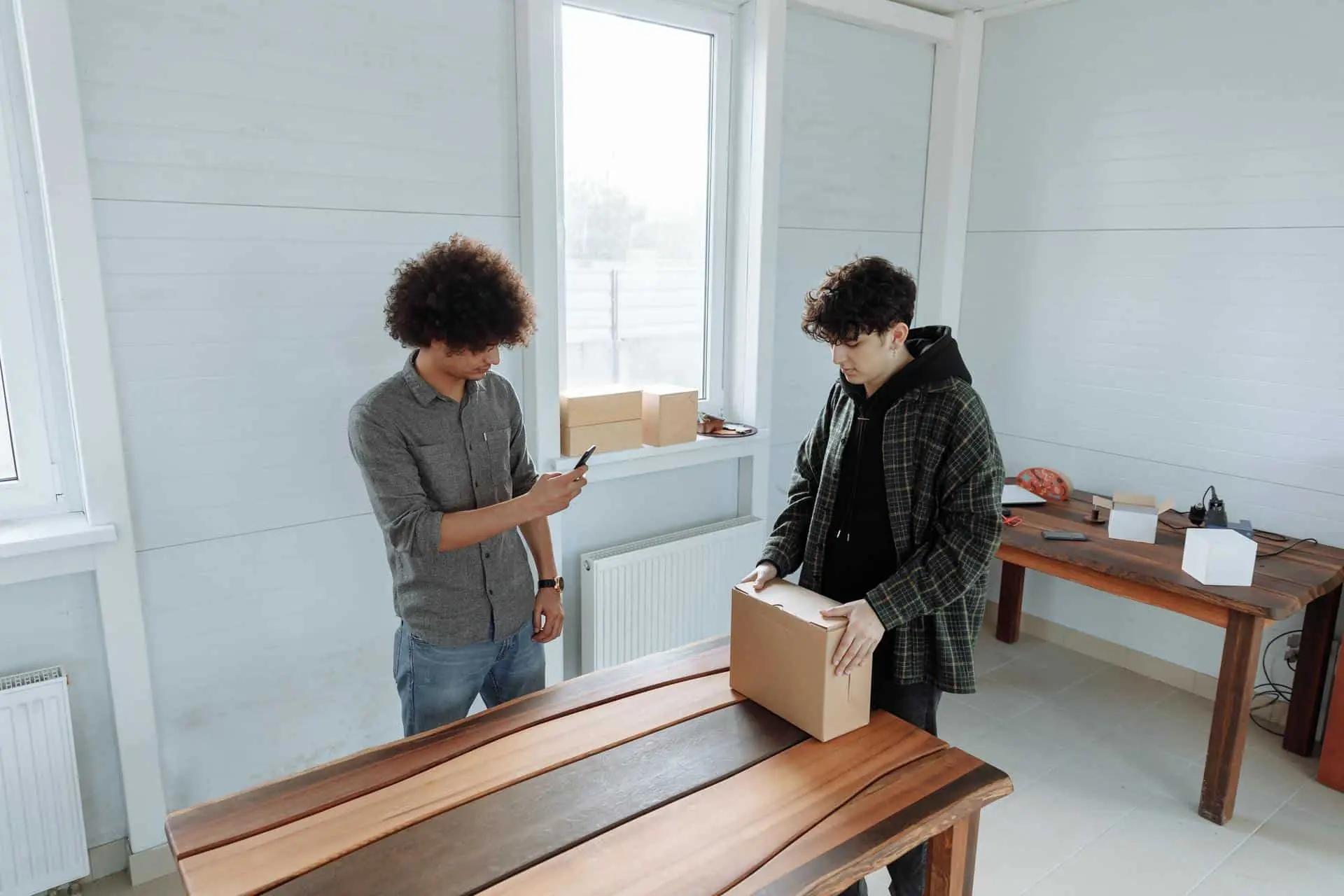 Two men in a workshop, one photographing a cardboard box on a wooden table with a phone