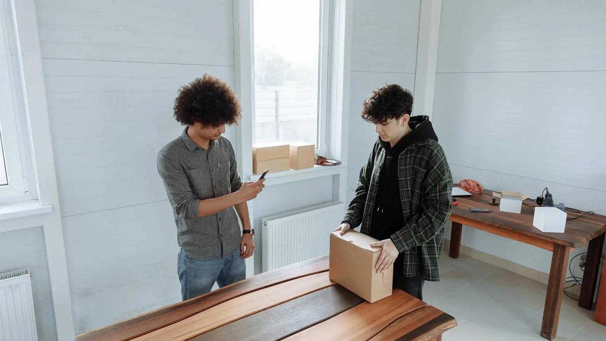 Two men in a workshop, one photographing a cardboard box on a wooden table with a phone