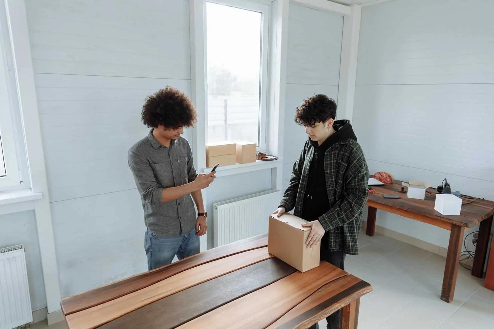 Two men in a workshop, one photographing a cardboard box on a wooden table with a phone