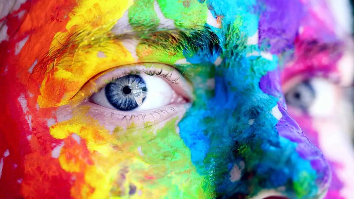 Close up of a person's face painted with bright, vibrant rainbow colors. Focus on a blue eye.