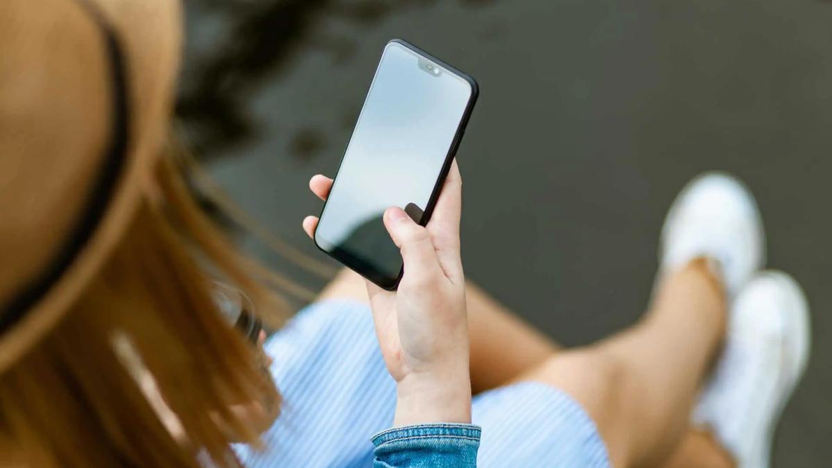Person in a straw hat, blue and white striped dress, and white sneakers holding a black smartphone with a blank screen