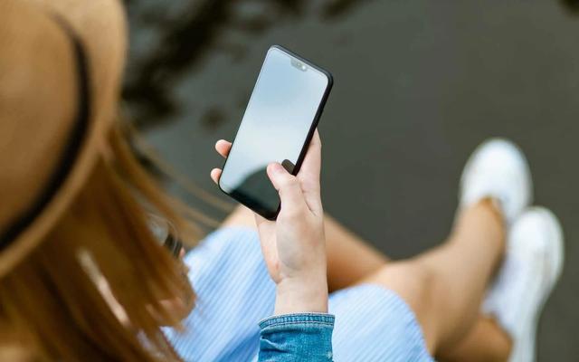 Persona con sombrero de paja, vestido de rayas azules y blancas, y zapatillas blancas sosteniendo un smartphone negro con pantalla en blanco