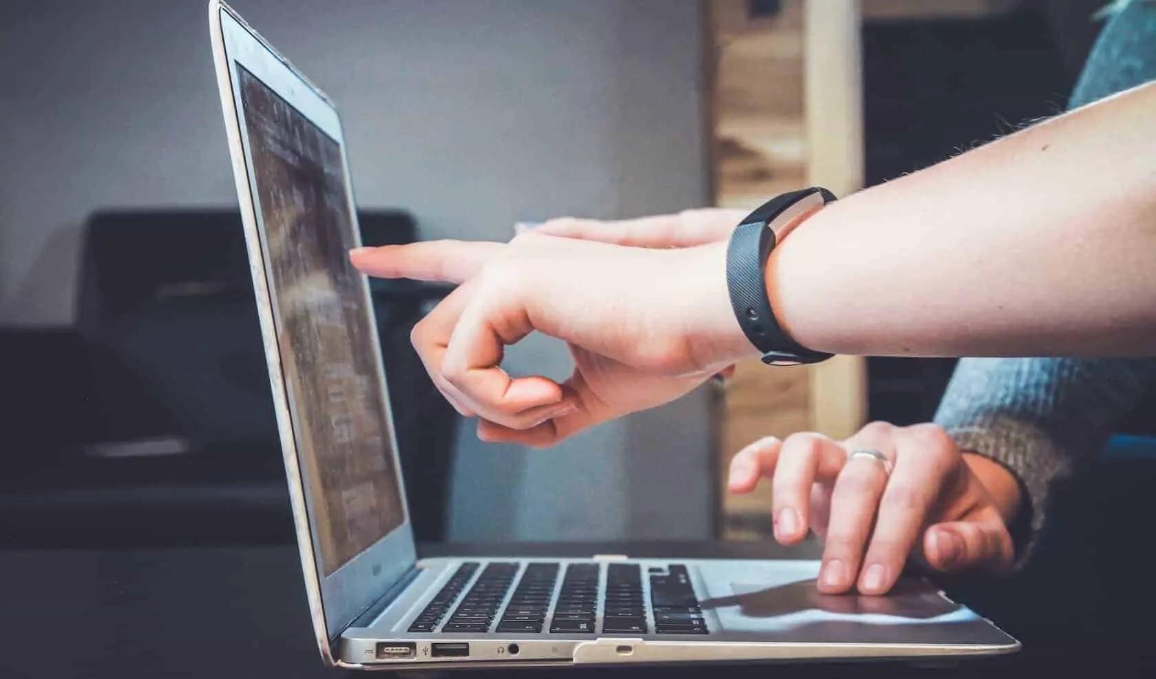 Una persona con pulsera de actividad señala la pantalla de un portátil mientras otra mano descansa en el panel táctil
