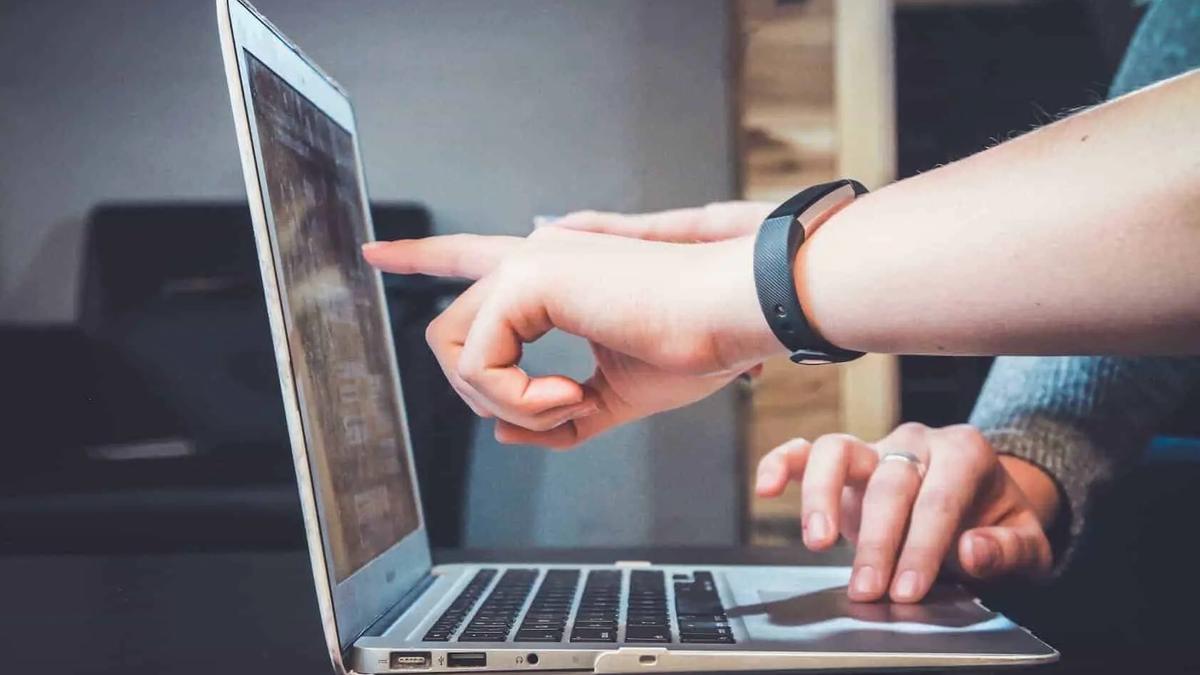 A person wearing a fitness tracker points at a laptop screen while another hand rests on the trackpad