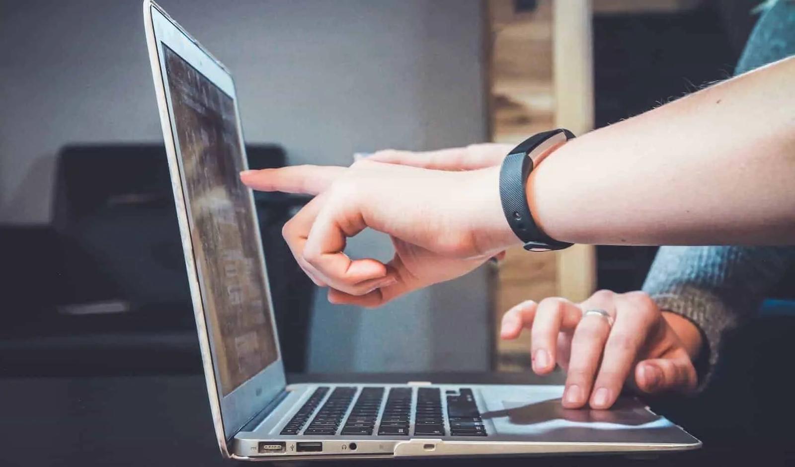 A person wearing a fitness tracker points at a laptop screen while another hand rests on the trackpad