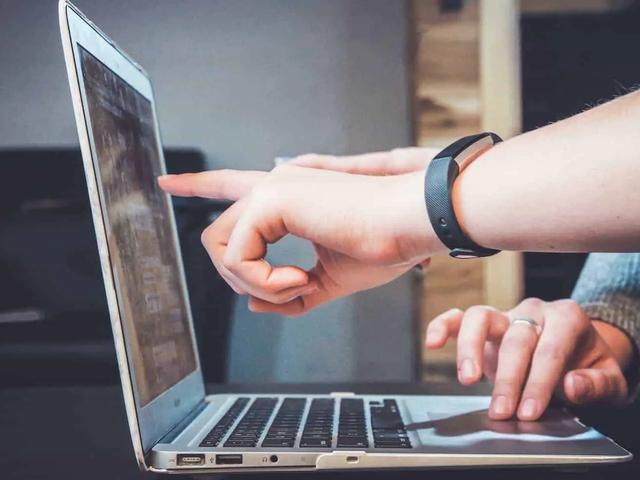 Una persona con pulsera de actividad señala la pantalla de un portátil mientras otra mano descansa en el panel táctil