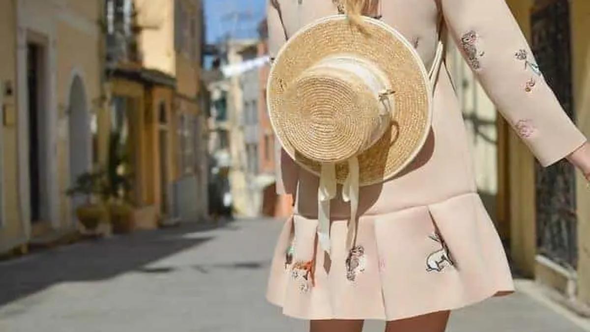 Woman in a pink patterned dress carrying a straw hat, walking down a narrow street