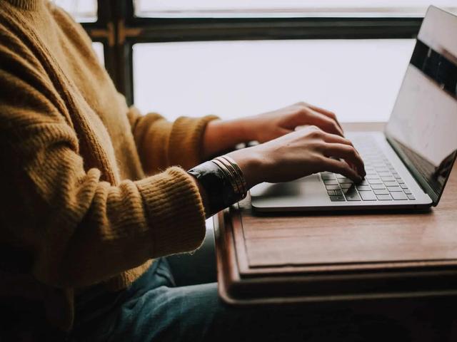 Person in a brown sweater and blue jeans typing on a silver laptop on a wooden table, window in background