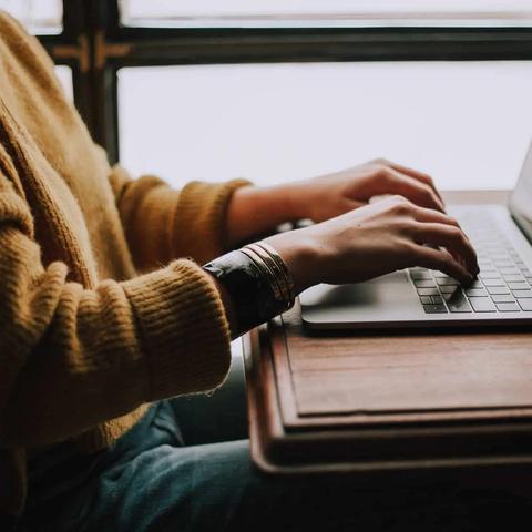 Person in a brown sweater and blue jeans typing on a silver laptop on a wooden table, window in background