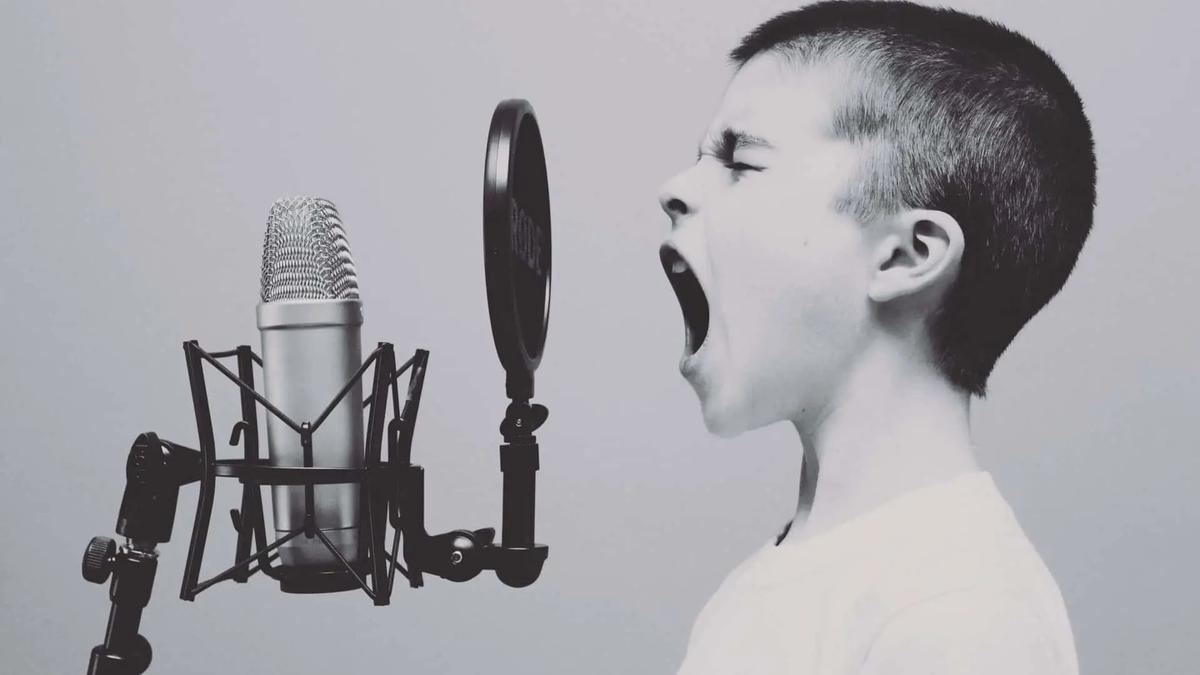 Black and white, profile of a young boy with mouth wide open, shouting into a studio microphone with a pop filter.