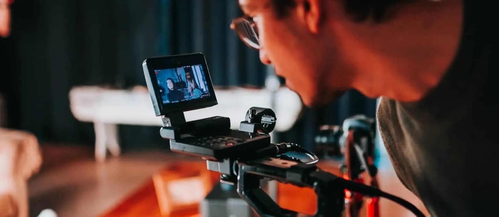 A person wearing glasses looks closely at a small camera monitor screen while filming