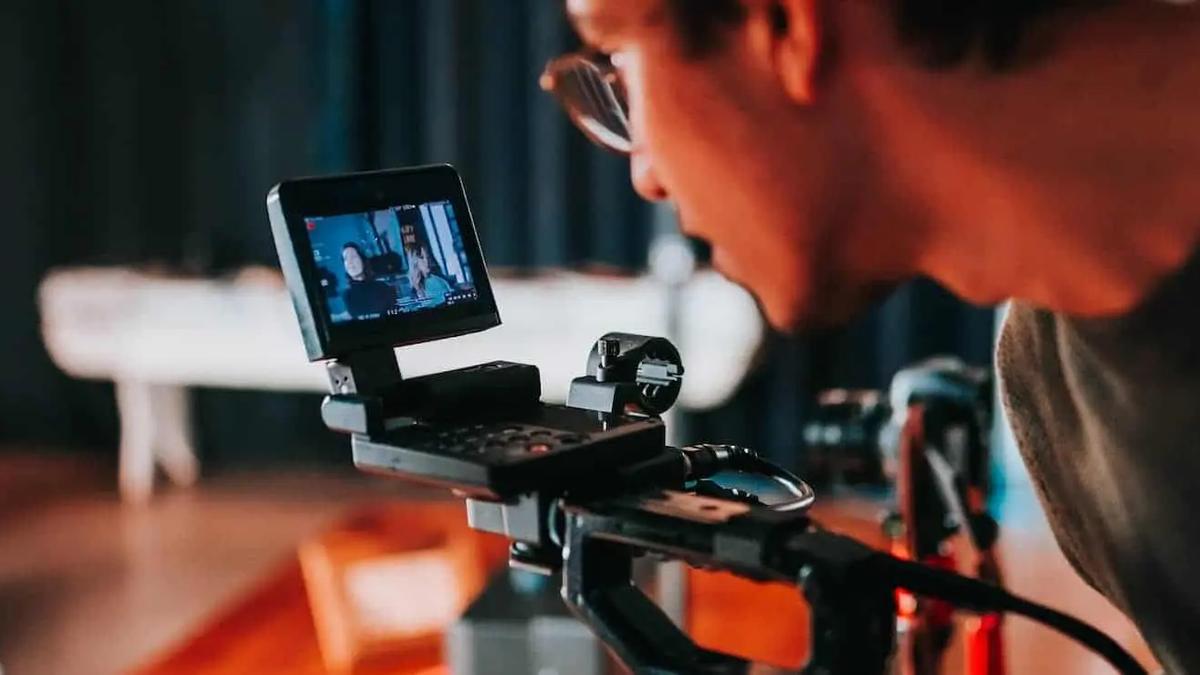 A person wearing glasses looks closely at a small camera monitor screen while filming