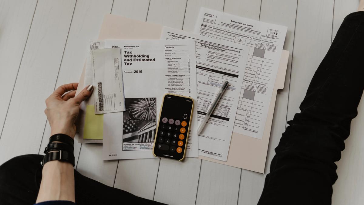 A person sits on the floor sorting tax documents and forms with a phone calculator and pen nearby