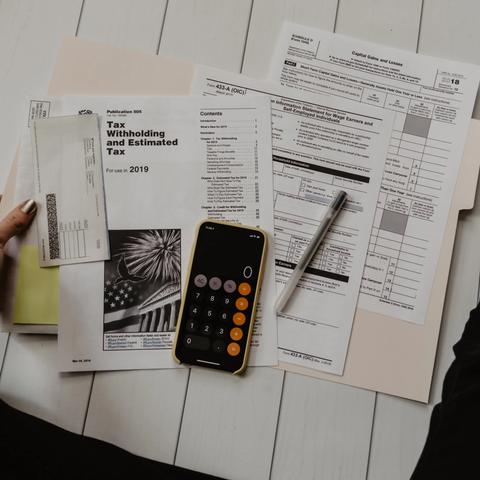 A person sits on the floor sorting tax documents and forms with a phone calculator and pen nearby