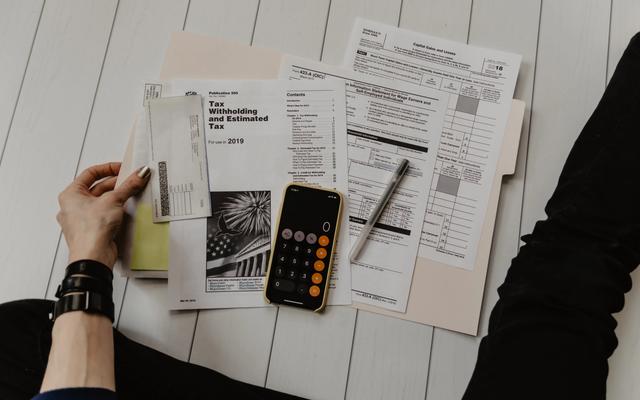 Una persona sentada en el suelo revisa documentos y formularios fiscales junto a un teléfono con calculadora y un bolígrafo