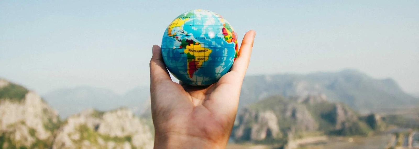 A hand holding a small toy globe against a mountain landscape