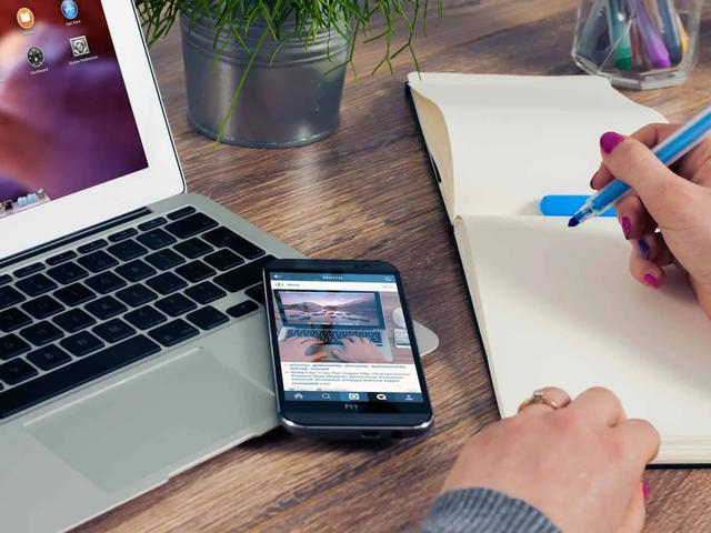 A phone showing an Instagram photo post sits beside a laptop while a hand writes in a notebook