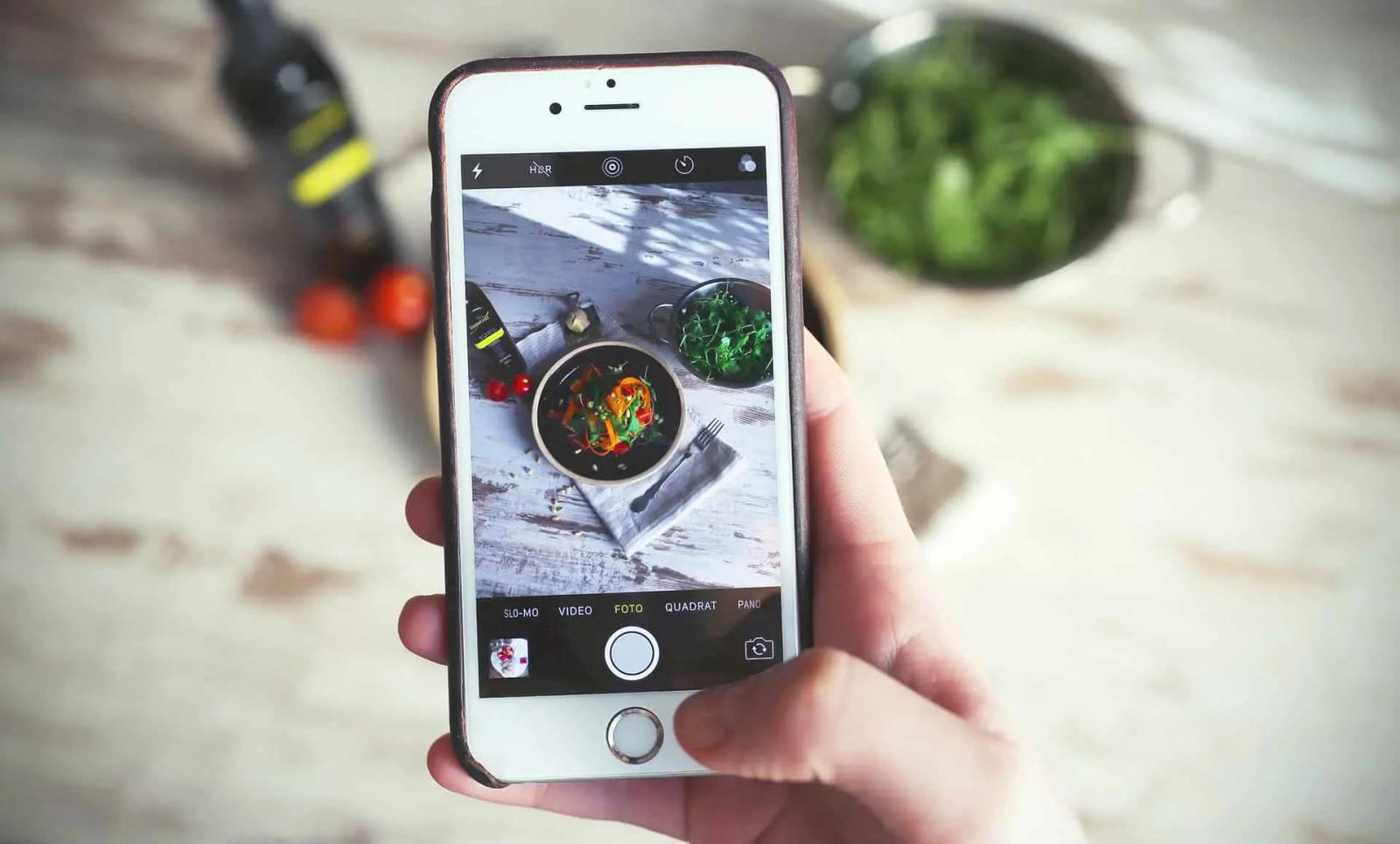 Una mano sostiene un telefono fotografiando una ensalada servida sobre una mesa de madera