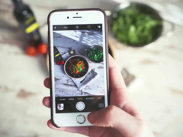 Una mano sostiene un telefono fotografiando una ensalada servida sobre una mesa de madera