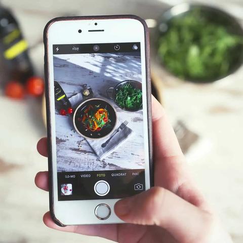 Una mano sostiene un telefono fotografiando una ensalada servida sobre una mesa de madera